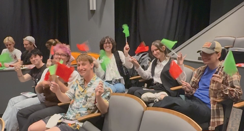 A group of students and CHEW educators sitting in an auditorium wave green flags and smile